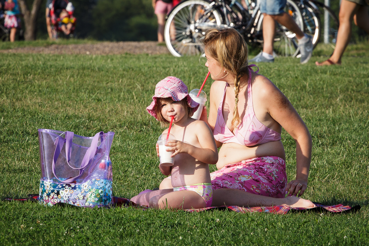 A woman and a child sit on a picnic blanket in a grassy area during Piens Fest 2014. Both are wearing pink swimwear and sun hats, drinking from straws. A transparent purple bag with items inside sits near them. Several people and bicycles are present in the background, enhancing the bright and sunny scene.