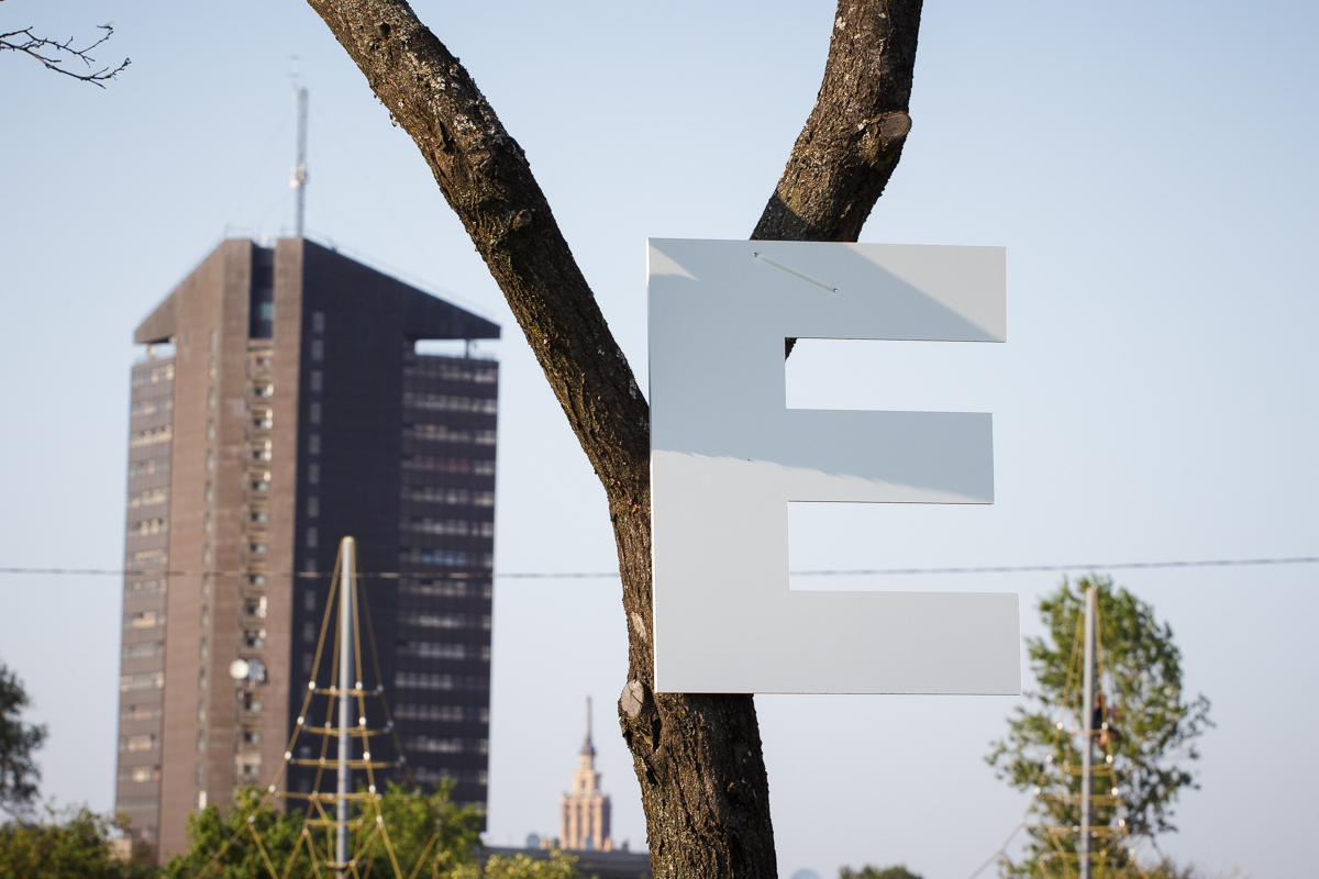A large, white, capital letter "E" is attached to the trunk of a leafless tree. In the background, there is a tall, brown, rectangular building with many windows, set against a clear blue sky at Piens Fest 2014. The lower part of the image contains some green foliage and the top of another building.