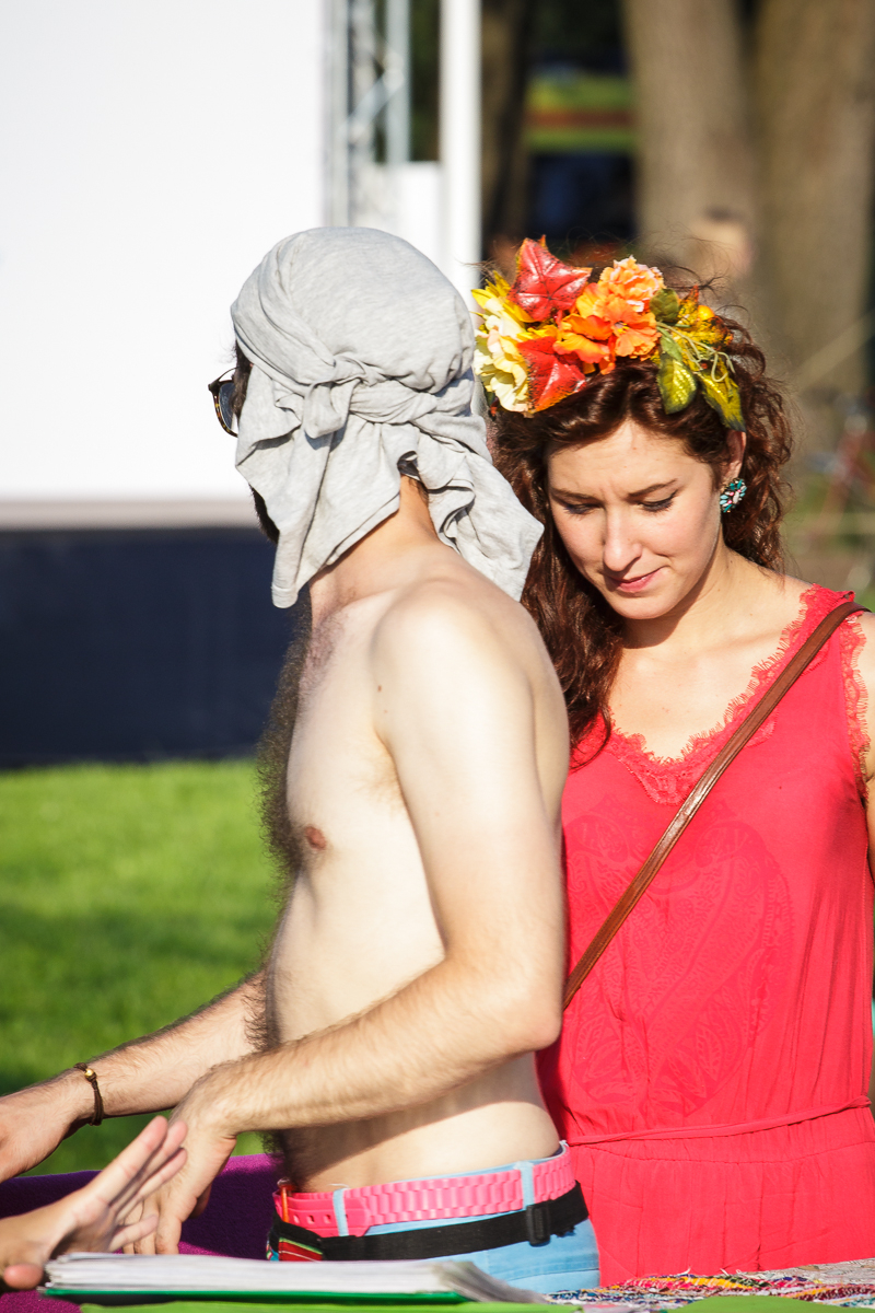 A shirtless person with a headscarf and sunglasses stands next to a woman dressed in a bright red dress and adorned with a vibrant flower crown. It's Piens Fest 2014, set outdoors on a sunny day, with grassy areas and trees in the background. The woman's gaze is downward, focused on something out of frame.