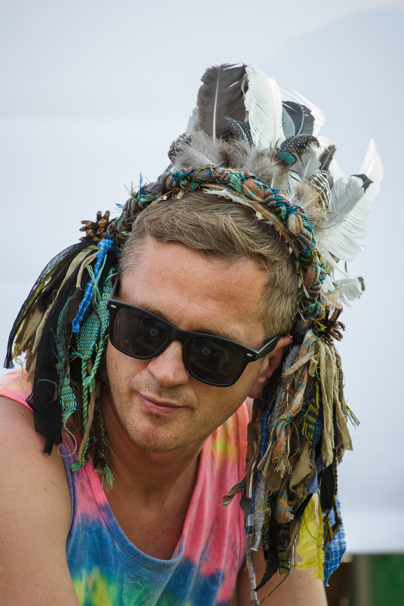 A man with short blonde hair wears black sunglasses and a headdress adorned with various colored feathers and cloth strips. He is dressed in a tie-dye tank top featuring shades of pink, yellow, and blue. The bright, blurred outdoor setting evokes memories of Piens Fest 2014.