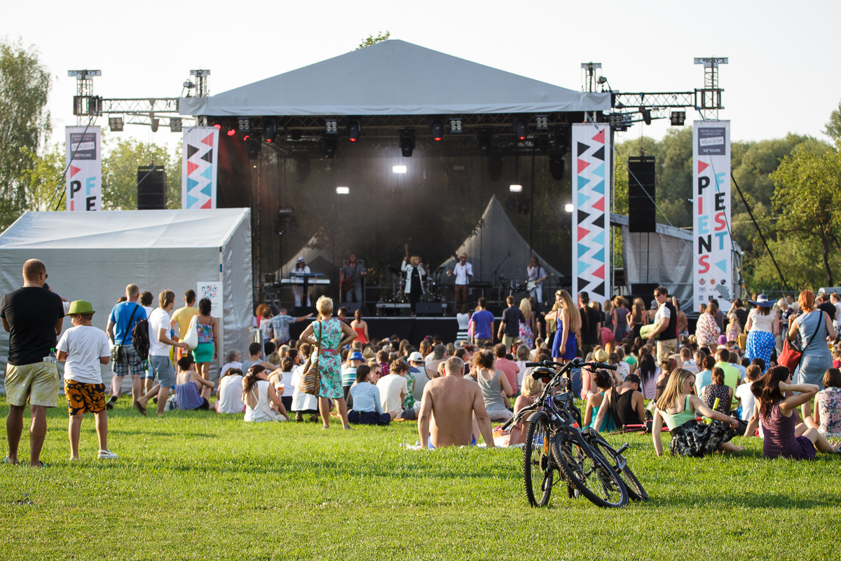 People gather at Piens Fest 2014, an outdoor music festival with a stage in the background. Some are sitting on the grass, while others stand. Bicycles are parked in the foreground. Stacks of speakers and colorful banners hang beside the stage, which is covered by a tent. Trees are visible in the distance.
