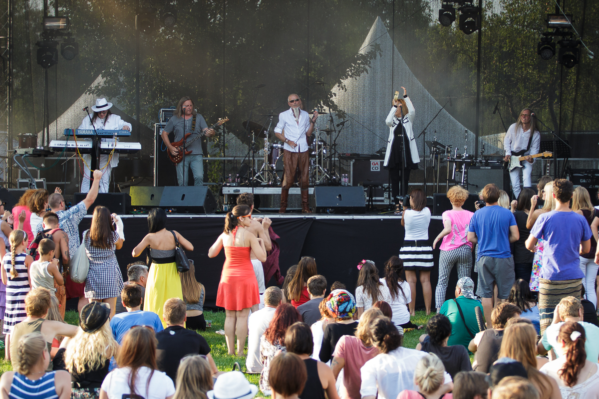 At Piens Fest 2014, a band performs live on an outdoor stage, featuring six musicians playing various instruments. An audience of diverse people stand and sit on the grass, watching enthusiastically. The stage is set against trees and is equipped with sound and lighting equipment, enhancing the lively atmosphere.
