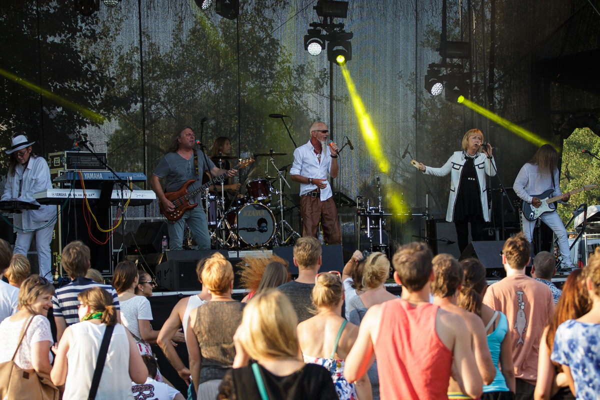 At Piens Fest 2014, a band performs on an outdoor stage with a drum set, keyboard, electric guitar, and bassist. Two vocalists sing at center stage, one dressed in a long jacket. Audience members, many in summer attire, watch and dance as sunlight filters through the trees, illuminating the scene.