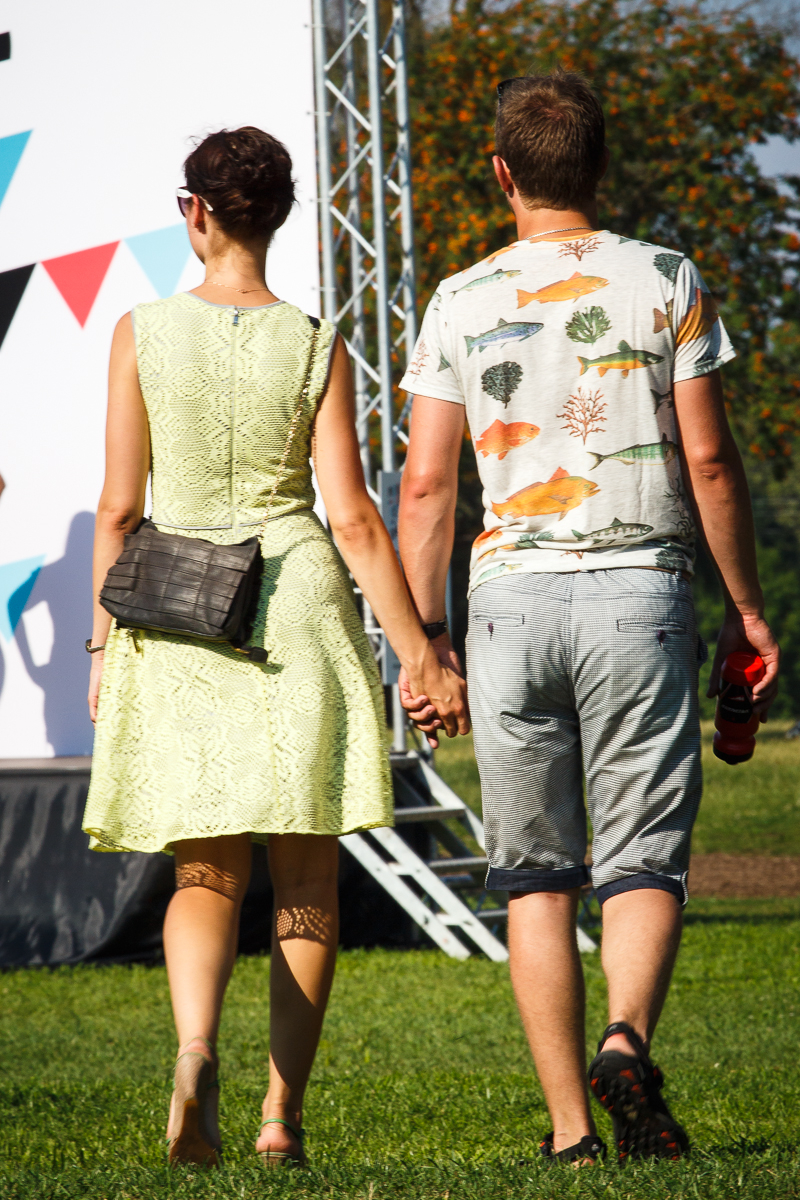 A couple is walking hand in hand in an outdoor setting during Piens Fest 2014. The woman is wearing a light green dress, sunglasses, and a black crossbody bag. The man is dressed in a white t-shirt with a colorful print and gray shorts, holding a red bottle. They are seen from behind on a sunny day.