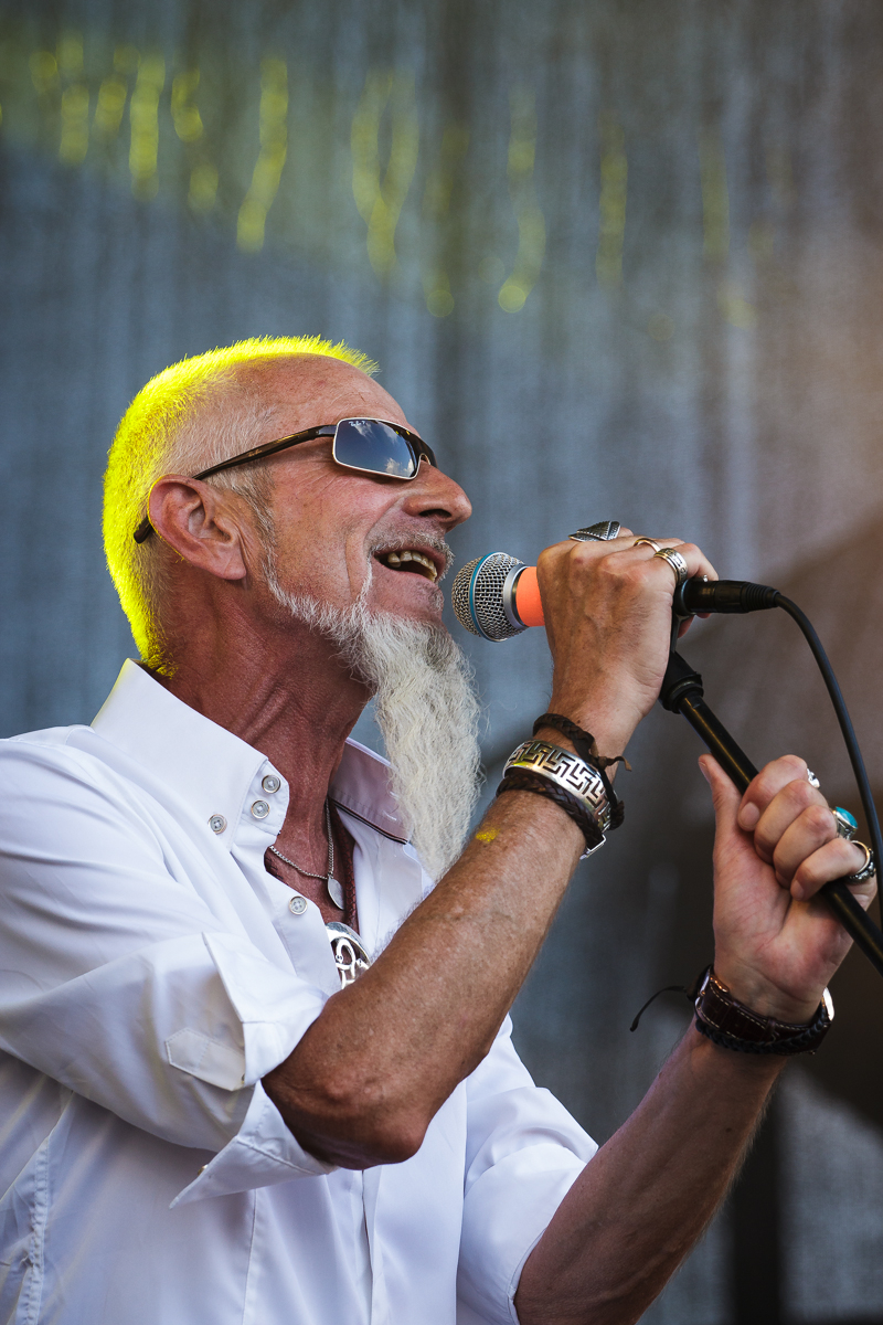An older man with a white beard, sunglasses, and a short-sleeved white shirt sings passionately into a microphone at the 2014 Piens Fest. He wears various bracelets, rings, and a necklace. The background, slightly out of focus, includes a mesh-like backdrop and yellow stage lighting.