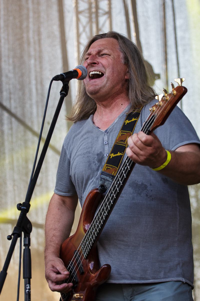 A man with long gray hair passionately sings into a microphone while playing a brown electric bass guitar on stage at Piens Fest 2014. He is wearing a gray short-sleeve shirt and a yellow wristband, with a "Fender" brand strap visible. The background features a mesh screen with stage equipment partially visible.