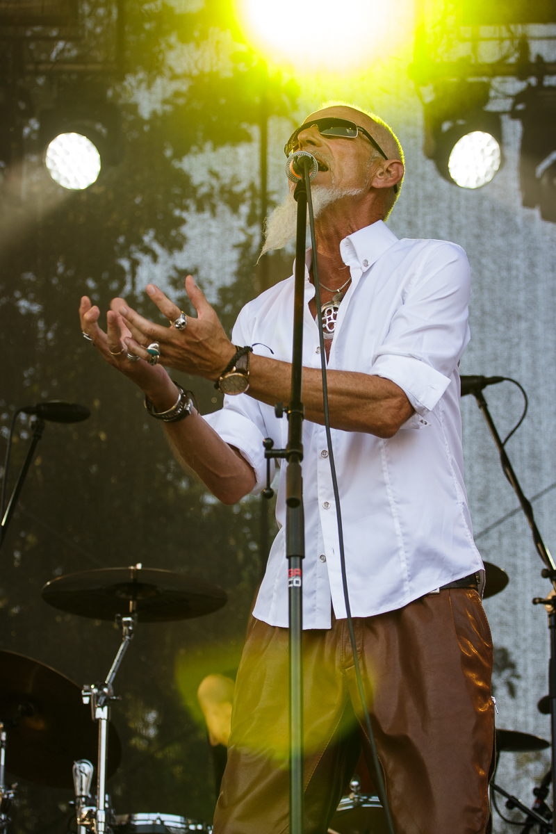 An elderly man with a white beard and sunglasses passionately sings into a microphone on the Piens Fest stage. He wears a white button-up shirt with rolled sleeves and brown pants, accessorized with rings, bracelets, and a necklace. Bright stage lights illuminate him while drum sets are visible in the background.