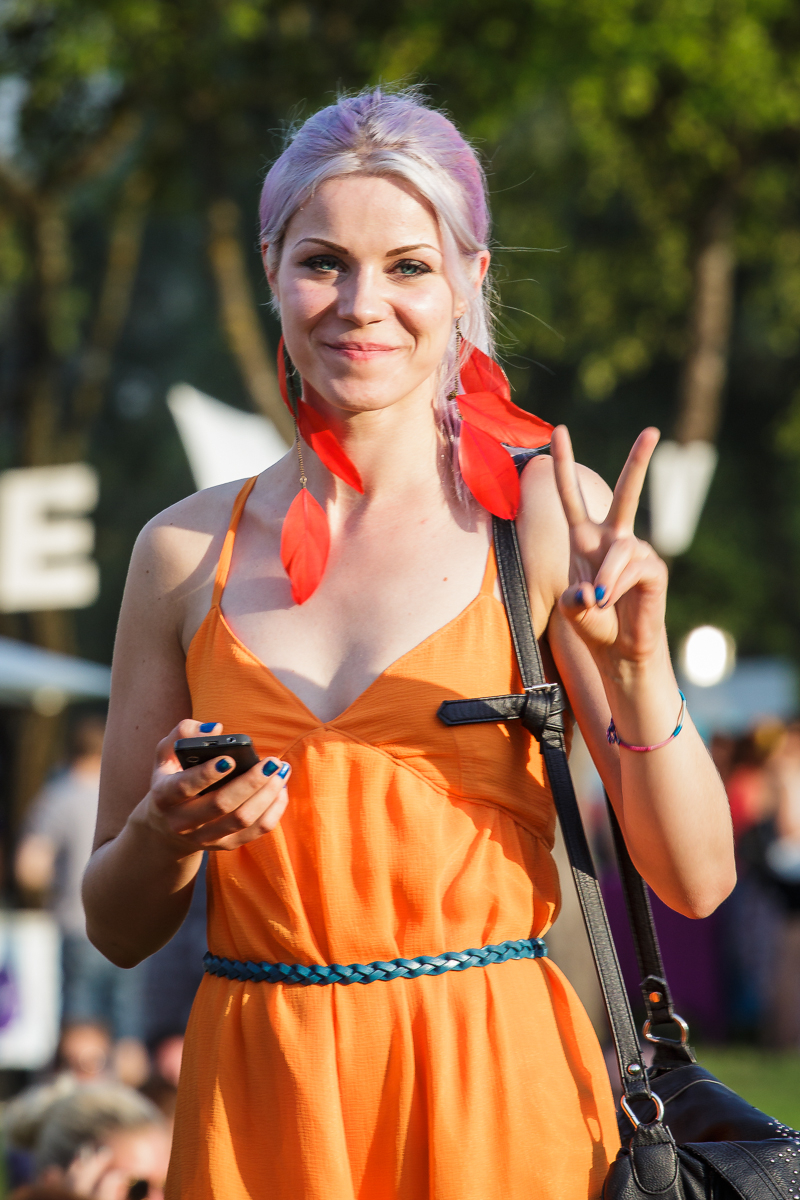 A woman with purple hair, wearing an orange dress, holds a phone and makes a peace sign with her right hand. She is accessorized with red feather earrings and a blue belt. The background is outdoors at the lively 2014 Piens Fest, filled with blurred greenery and people enjoying the festivities.
