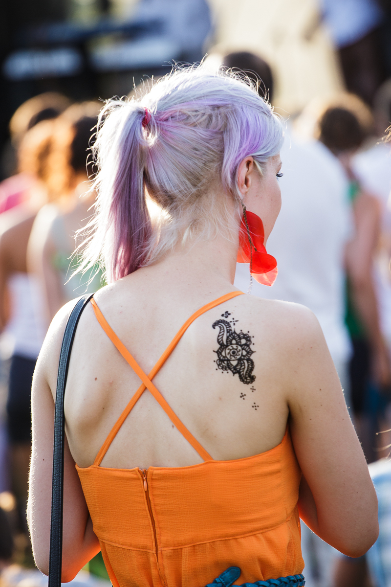 A woman with light purple hair pulled into a ponytail stands in a crowd at Piens Fest 2014. She is wearing a bright orange dress with thin straps crossing over her back. On her left shoulder blade is a large, intricate black henna tattoo. Her left earring is large and red. She has a black strap over her shoulder.