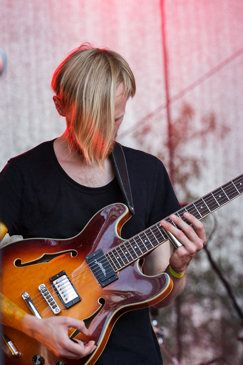 A person with blond hair, dressed in a black shirt, is playing an amber-colored electric guitar on stage at Piens Fest 2014. Focused on the instrument, the guitar strap sits over their shoulder. The background is a textured, light-colored curtain under red stage lighting.