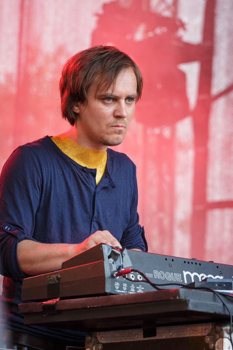A person with medium-length brown hair, wearing a blue shirt with yellow detail at the collar, intensely plays a keyboard adorned with "Rogue" and "Moog" labels. The red and blurred background, coupled with an indistinct shadowy figure, suggests this could be a live performance at Piens Fest 2014.