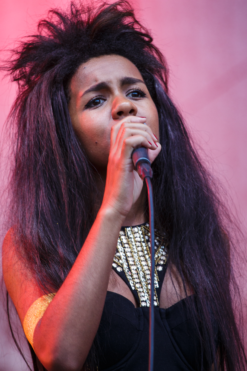 A woman with long, voluminous hair sings into a microphone at Piens Fest 2014. She wears a black strapless dress adorned with a gold beaded neckline and a gold armband. The background is out-of-focus, pink, and gradually transitions to white. Her facial expression is focused and passionate.