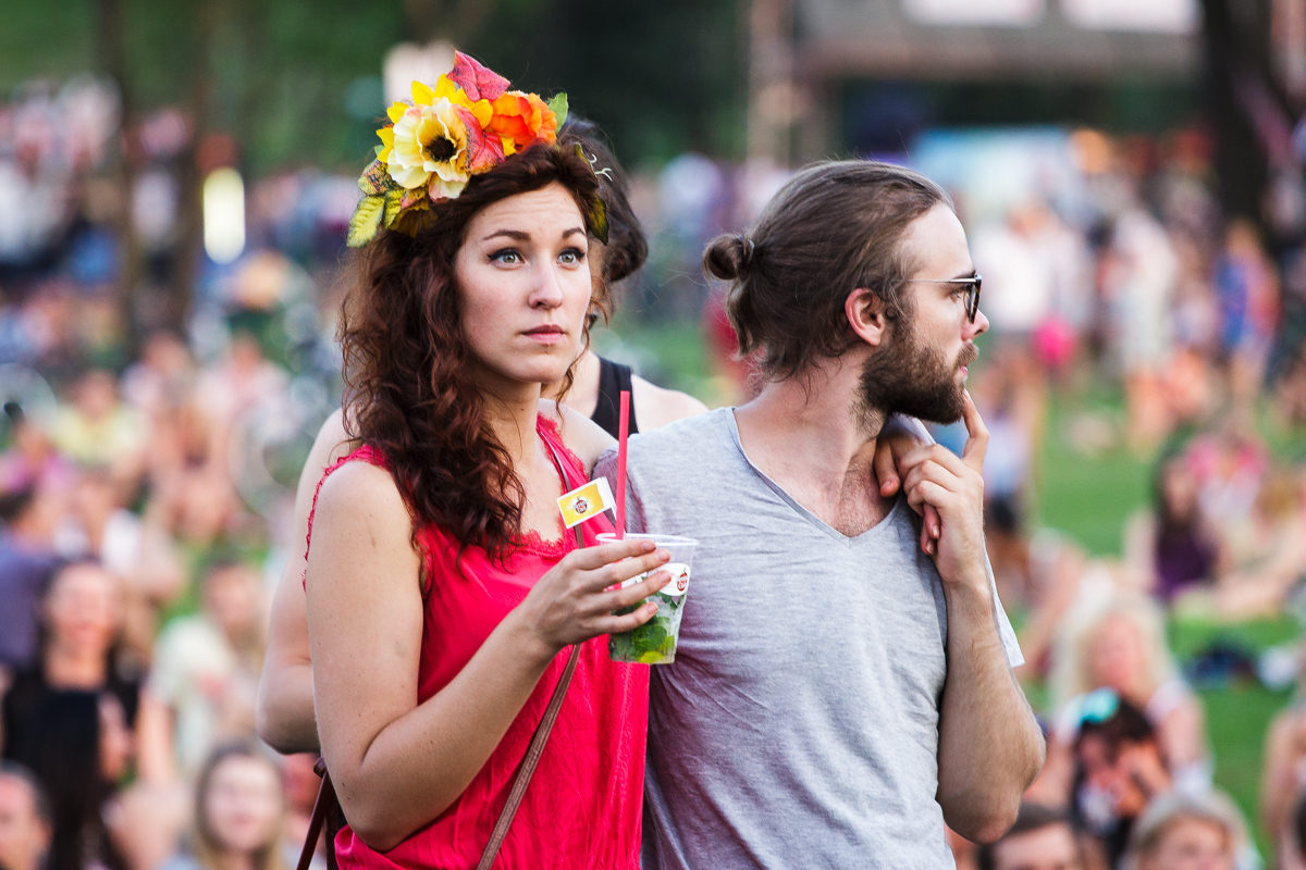 A woman wearing a floral headpiece and a pink dress holds a clear cup while standing beside a man with a beard, sunglasses, and a gray T-shirt. The man is looking to his left with a cigarette in his mouth. The background shows people sitting and standing on the grassy area of Piens Fest 2014, indicating an outdoor event.