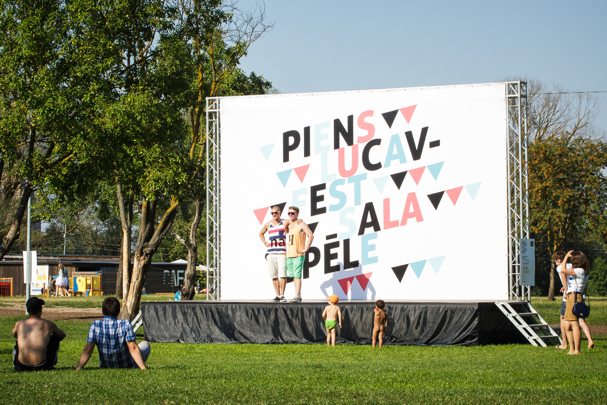 Two people stand on an outdoor stage beside a large sign with colorful, geometric text at Piens Fest 2014. They pose for a photo on a sunny day in the park. Several people sit or lie on the grass, enjoying the open, grassy field and warm weather. Trees and playground equipment are visible in the background.