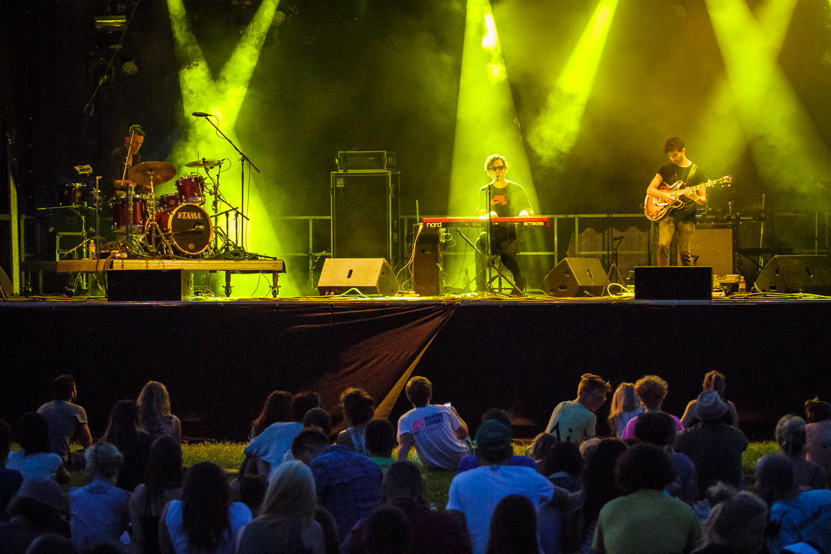 A live band performs on stage with dramatic yellow lighting at Piens Fest 2014. The drummer is on the left, a keyboard player in the middle, and a guitarist on the right. An audience sits on the grass, watching intently. Smoke effects and amplifiers are visible in the background.