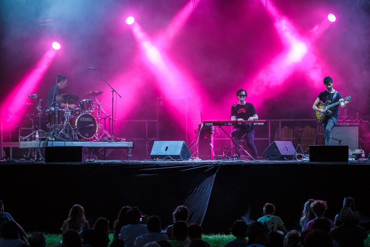 A live music performance on an outdoor stage with three musicians at Piens Fest 2014. From left to right, a drummer, a keyboardist, and a guitarist play under vibrant pink and purple stage lights, producing a misty, atmospheric effect. An audience watches attentively from the grass in front of the stage.