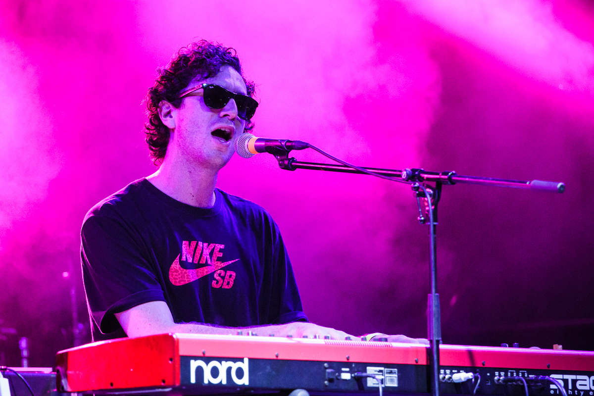 A person with curly hair, wearing sunglasses and a black Nike SB shirt, sings into a microphone while playing a red and white Nord keyboard on stage at Piens Fest 2014. The background is lit with purple and pink lights, creating a vibrant atmosphere.
