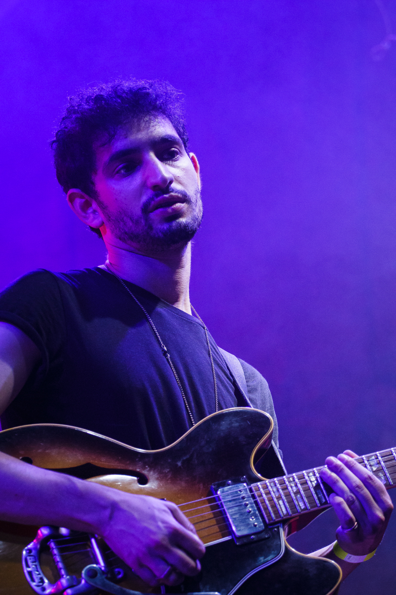 At Piens Fest 2014, a man wearing a black T-shirt plays an electric guitar on stage. He has short curly hair and a beard. The background is illuminated with purple lighting, creating a vibrant atmosphere. The man looks slightly upwards, engrossed in the music he is playing.