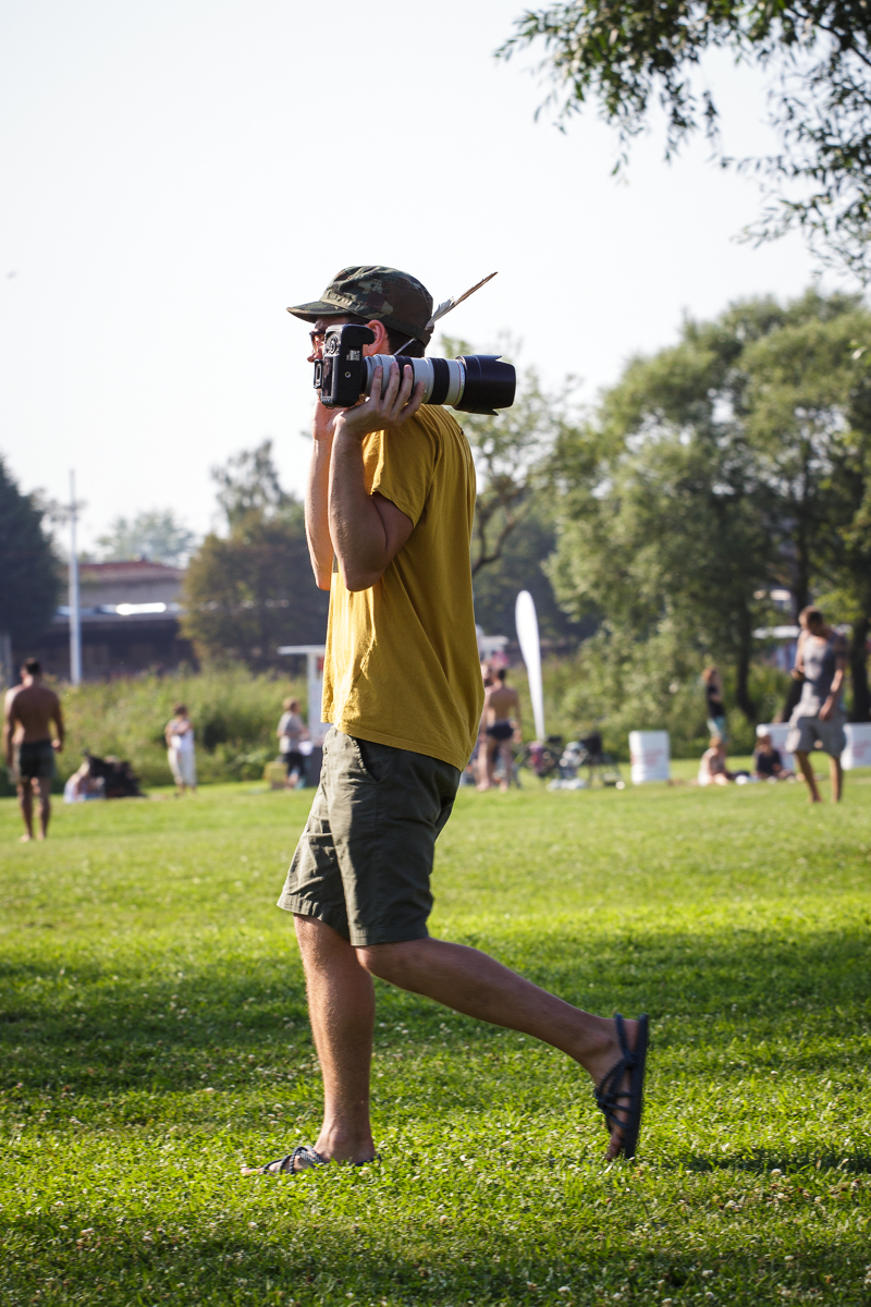 A person dressed in a yellow t-shirt, camouflage hat, shorts, and flip-flops is walking on a grassy field at Piens Fest 2014. They are carrying a camera with a long telephoto lens on their shoulder. The background shows other people relaxing and enjoying a sunny day in the park with trees and blue sky.