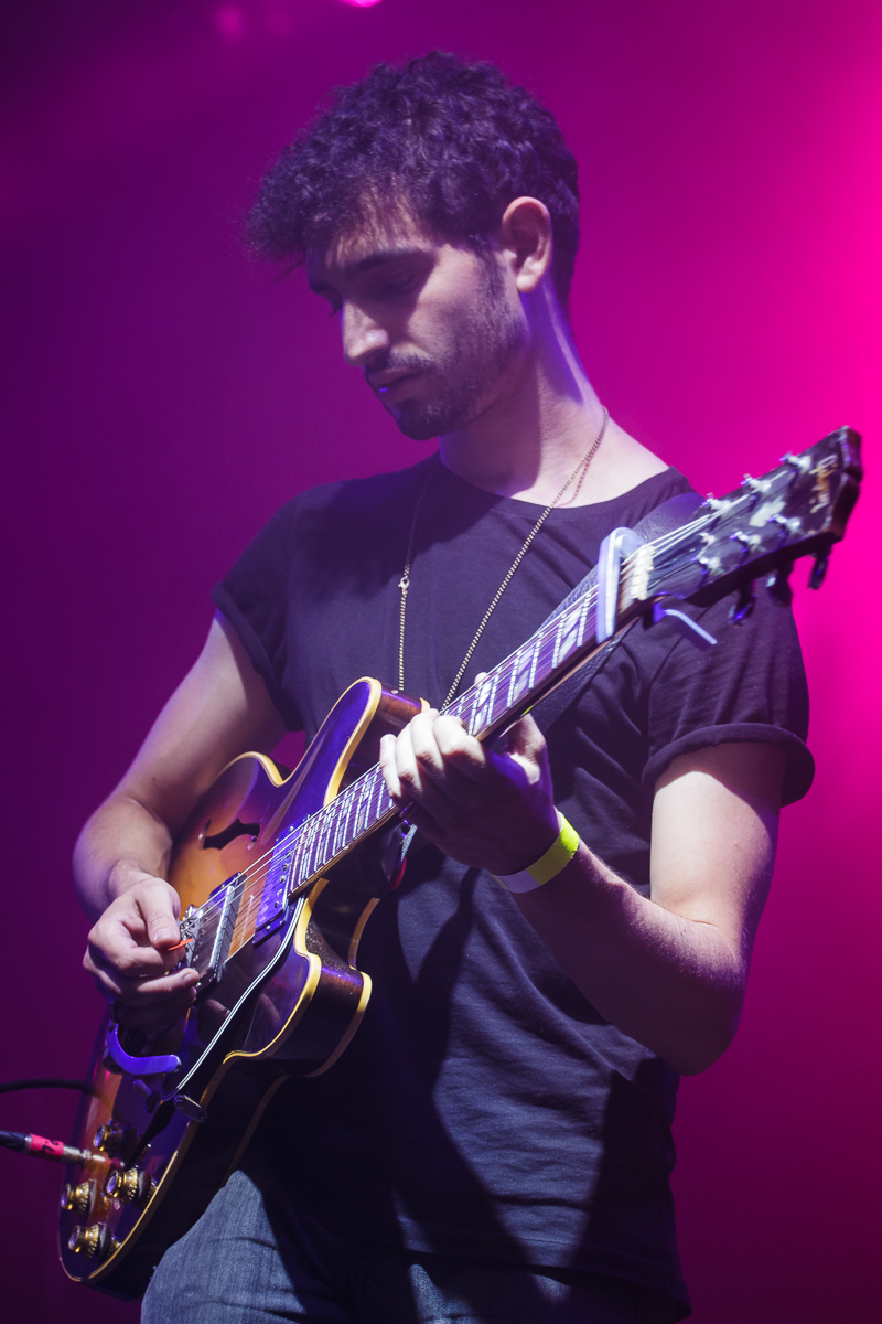 At Piens Fest 2014, a guitarist with dark, curly hair and a trimmed beard stands on stage, playing an electric guitar. He wears a black t-shirt and a yellow wristband on his right arm. The background is illuminated with purple and pink stage lights, creating a vibrant atmosphere.