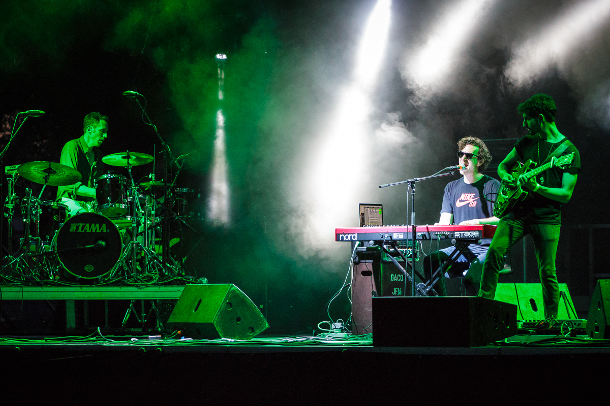 At Piens Fest 2014, a three-member band performs on stage. The drummer is playing a drum set on the left, the keyboardist is in the center singing into a microphone, and the guitarist stands on the right. Green and white stage lights and fog create a dramatic atmosphere. The drum kit has "TAMA" written on it.