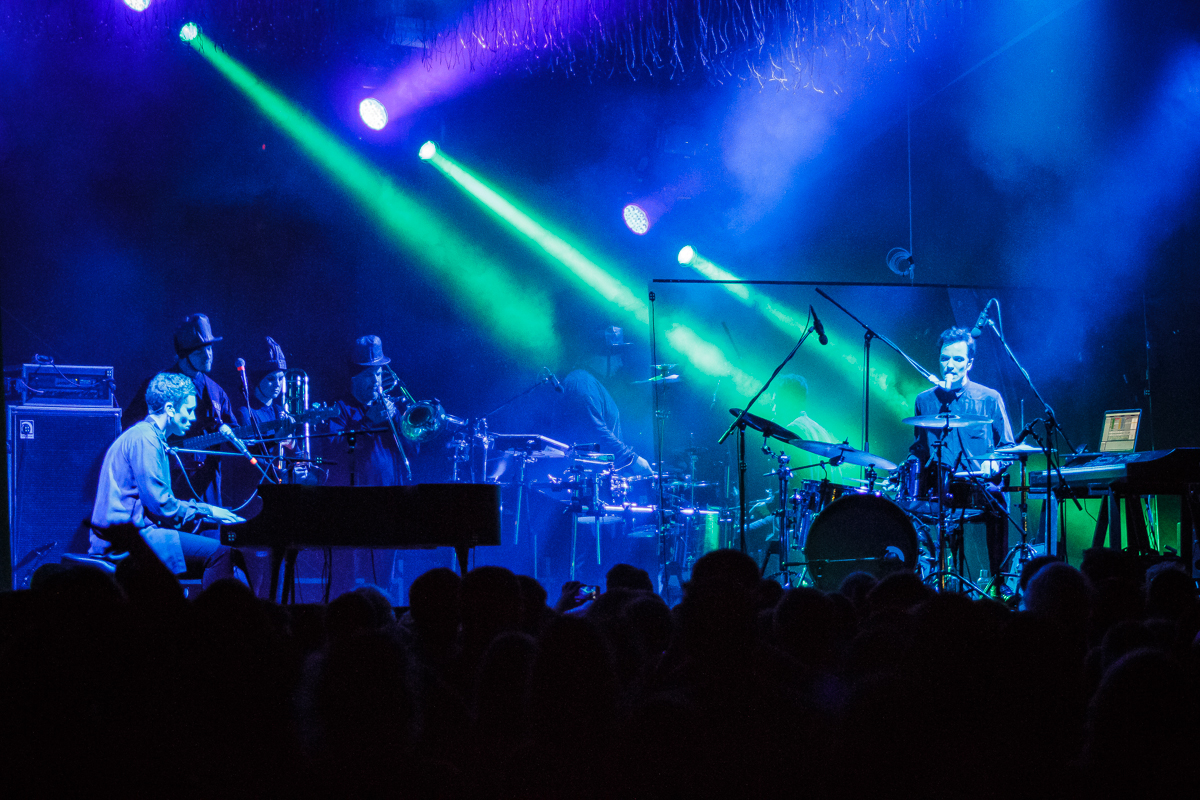 At Piens Fest 2014, a live music performance unfolded on a dimly lit stage. A pianist and drummer took the foreground while musicians playing brass instruments stood behind them, surrounded by sound equipment. Vibrant blue and green lights illuminated the stage as the captivated crowd watched.