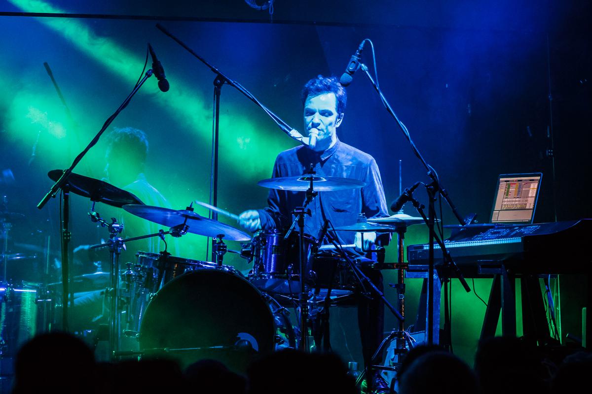A drummer sits at a drum set on stage during a live performance at Piens Fest, illuminated by vibrant blue and green stage lights. Several microphones surround the setup, and a keyboard is to the drummer’s right with a laptop screen visible. The background includes faint outlines of other band members from 2014.