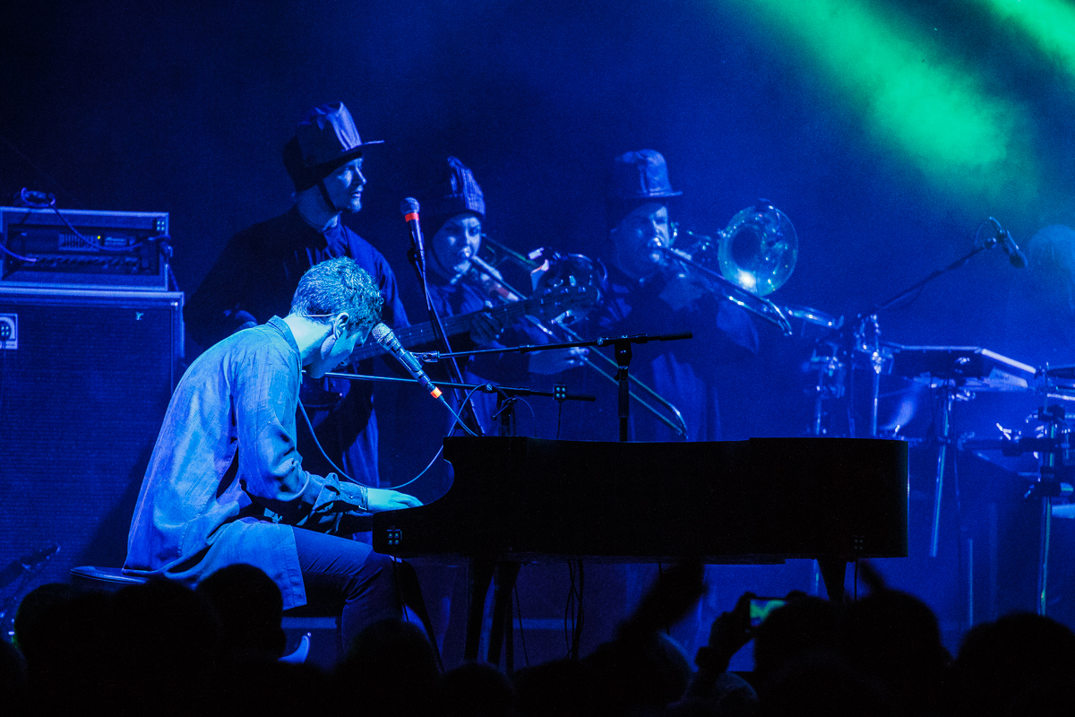 A musician plays a grand piano on stage under blue and green lights at Piens Fest 2014, while a brass ensemble in the background plays trombones and trumpets. The audience's silhouettes are visible in the foreground, and there is musical equipment to the left, suggesting an electric live performance.