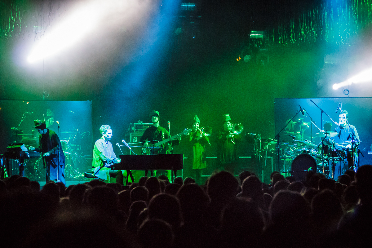 At Piens Fest 2014, a live band performs on stage with vibrant green and blue lighting. The ensemble includes a pianist, two brass players, a guitarist, bassist, drummer, and a vocalist. All wearing dark clothing, the band captivates the silhouetted audience in the foreground who are thoroughly enjoying the performance.