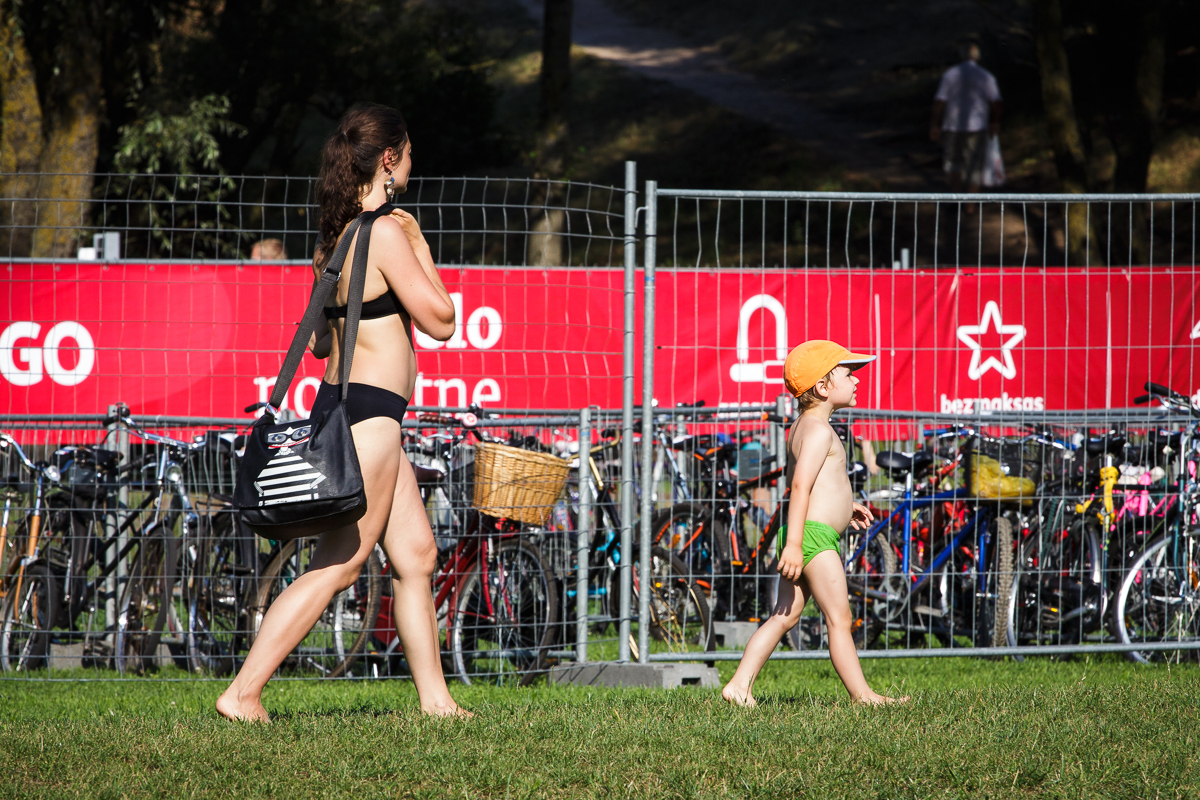A woman in a black bikini walks barefoot on grass, carrying a black shoulder bag, accompanied by a young child wearing green shorts and a yellow hat. Behind them, bicycles are parked behind a wire fence, and a red Piens Fest 2014 sign with white graphics stands tall.