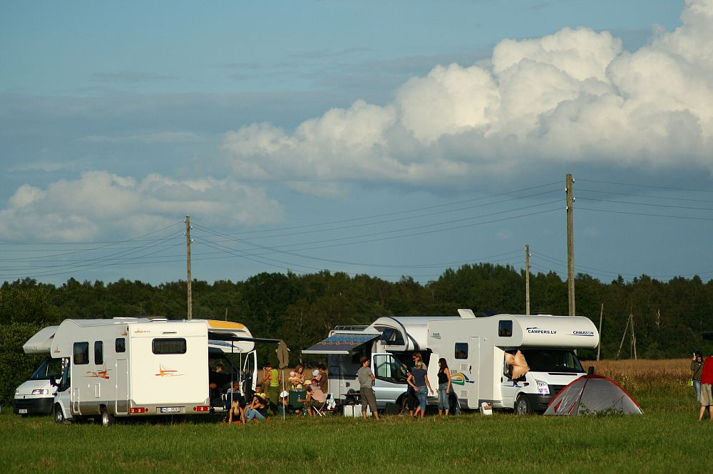 A group of people are enjoying a camping trip in a grassy field with two white RVs and a tent. They are sitting and standing around, engaging in conversation and activities reminiscent of Positivus AB 2008. The sky is partly cloudy, and there are trees and utility poles in the background.