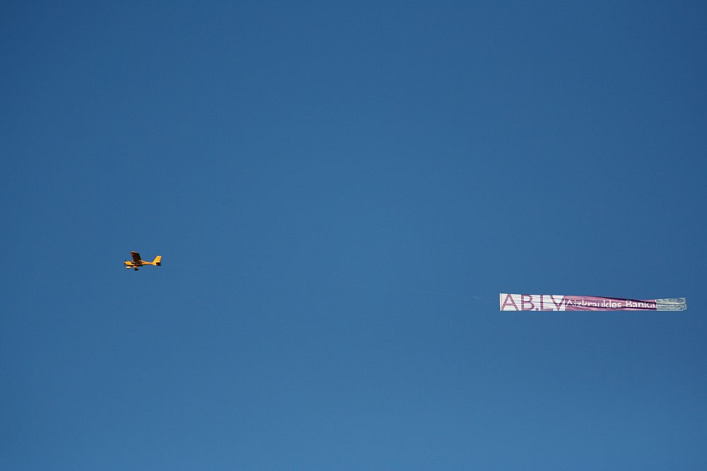 A small yellow airplane is flying in a clear blue sky, towing a long rectangular banner. The banner is partially readable, displaying the letters "ABLY" and some additional text that is not fully legible, possibly promoting Positivus 2008. The scene is set against a plain backdrop with no clouds visible.