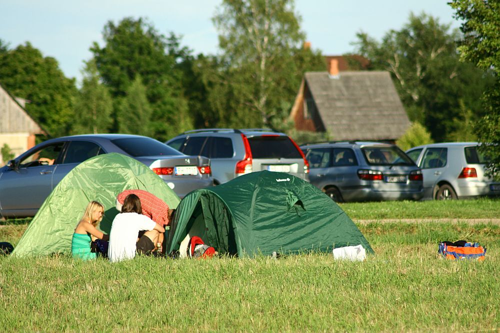Two green camping tents are set up on a grassy field. Two people sit on the grass near the tents, with one holding a red and white checkered cloth, reminiscent of a Positivus festival scene. Multiple parked cars are in the background, along with trees and houses under a clear sky.