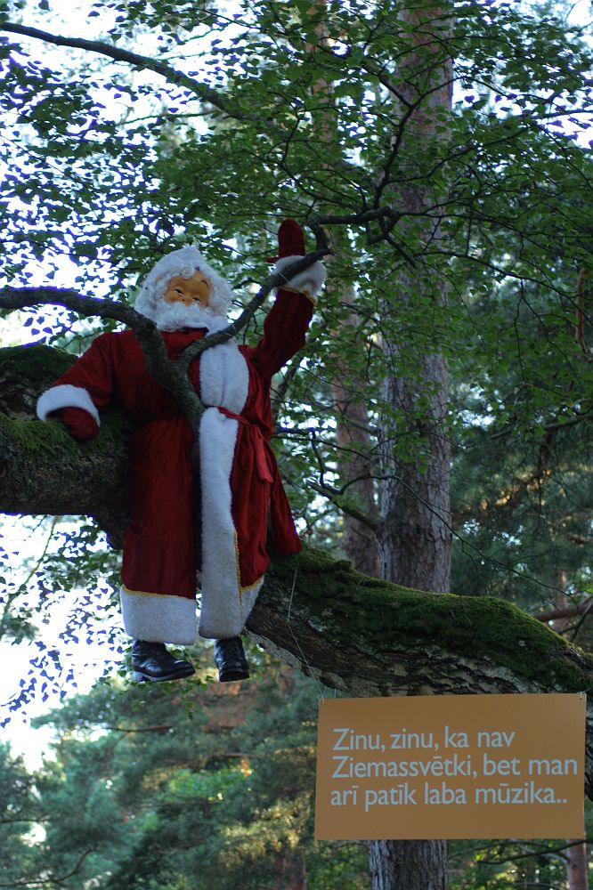 A Santa Claus figure in a red suit and white beard is sitting on a tree branch outdoors surrounded by greenery, playing a guitar. In the bottom right corner, there is a text box with Latvian text that reads, “Zinu, zinu, ka nav Ziemassvētki, bet man arī patīk laba mūzika..”. This scene feels like an AB 200