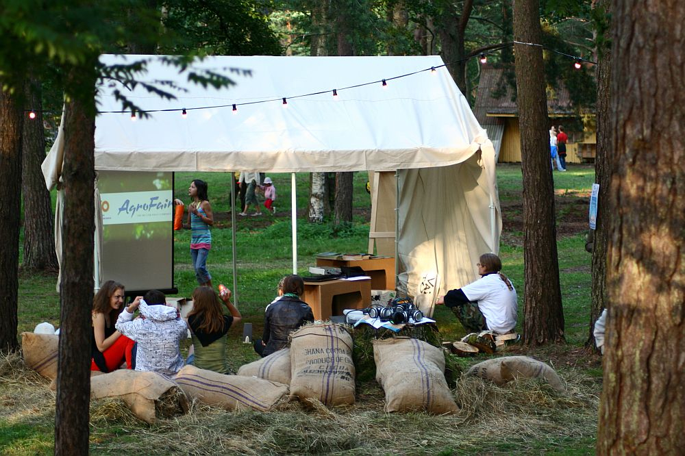 A small white tent is set up in a grassy forest clearing, surrounded by trees. People are sitting on hay bales and large sacks outside the tent, engaging in conversation. String lights are hung above the tent. In the background, more people are walking near various small buildings, reminiscent of Positivus AB 2008.