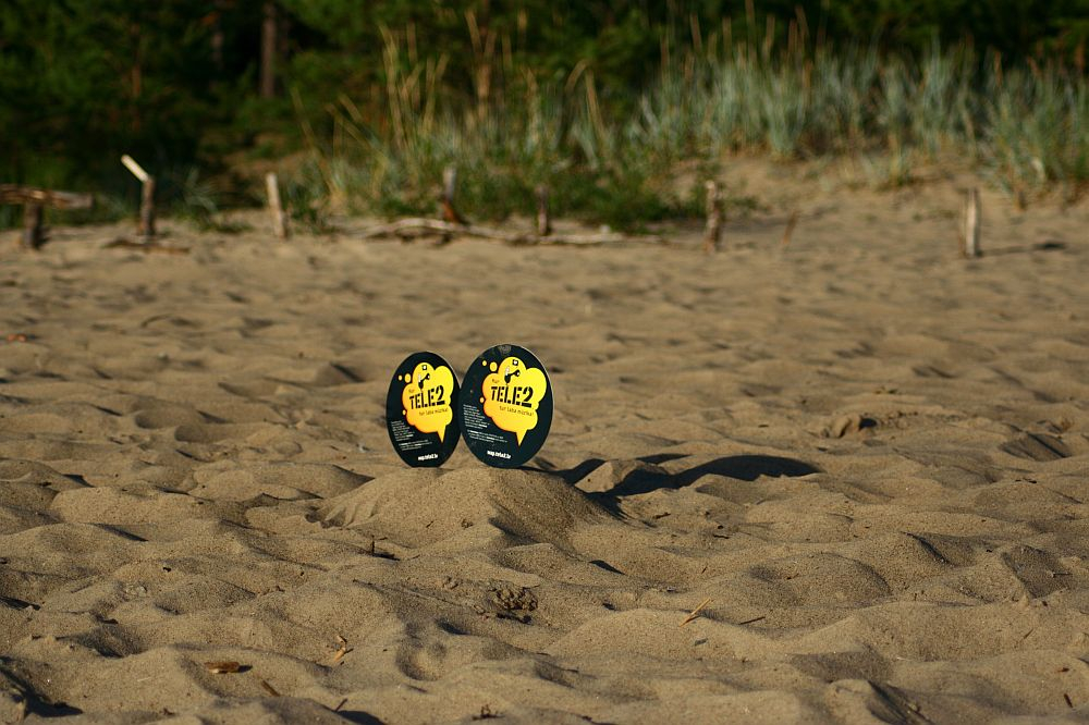 Two oval-shaped signs are placed in a sandy area, likely a beach, surrounded by dune grass and trees in the background. The signs have a yellow and black design with the text "tie. 2" prominently displayed. The sunny scene casts clear shadows of the signs on the sand, reminiscent of Positivus AB 2008 vibes.
