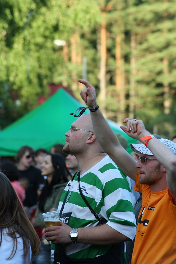 A person in a green and white striped shirt holds a cup while raising their other hand at the Positivus 2008 festival. Beside them, another attendee in an orange shirt and white hat gestures enthusiastically. In the background, trees and a green tent nestle among a lively crowd of AB festival-goers.