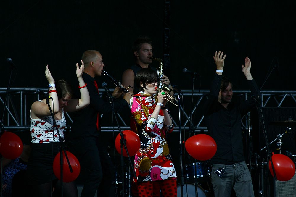At Positivus 2008, a live band performs on stage. The musicians, including a woman in a colorful dress playing the trumpet, are surrounded by red balloons. Three people in the foreground have their arms raised. The background features AB stage equipment and dim lighting.
