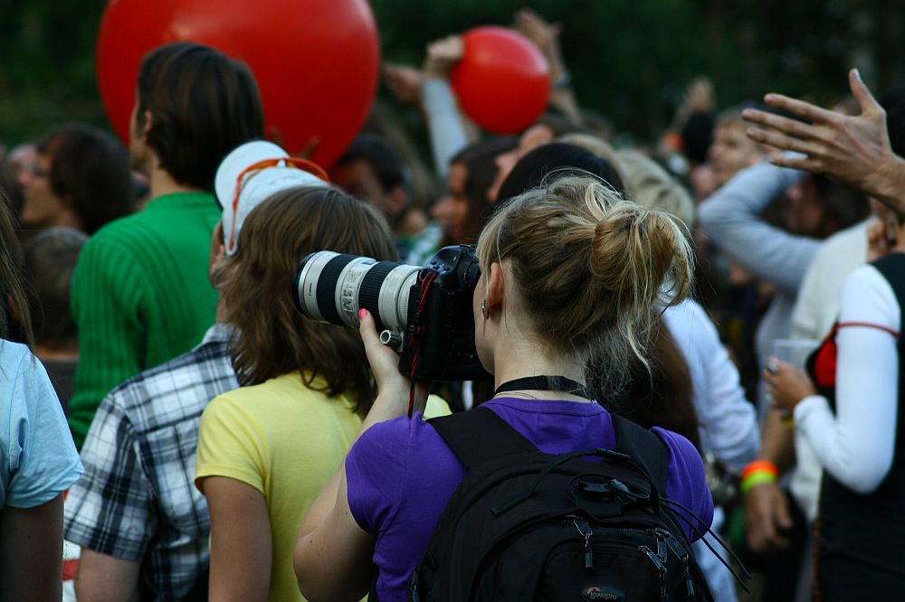 A woman with blond hair in a ponytail, wearing a purple shirt and black backpack, uses a large lens camera to photograph the Positivus festival. Around her, people hold up red balloons and raise their hands, some facing away from the camera as they immerse themselves in AB 2008's vibrant atmosphere. The background is slightly blurred.