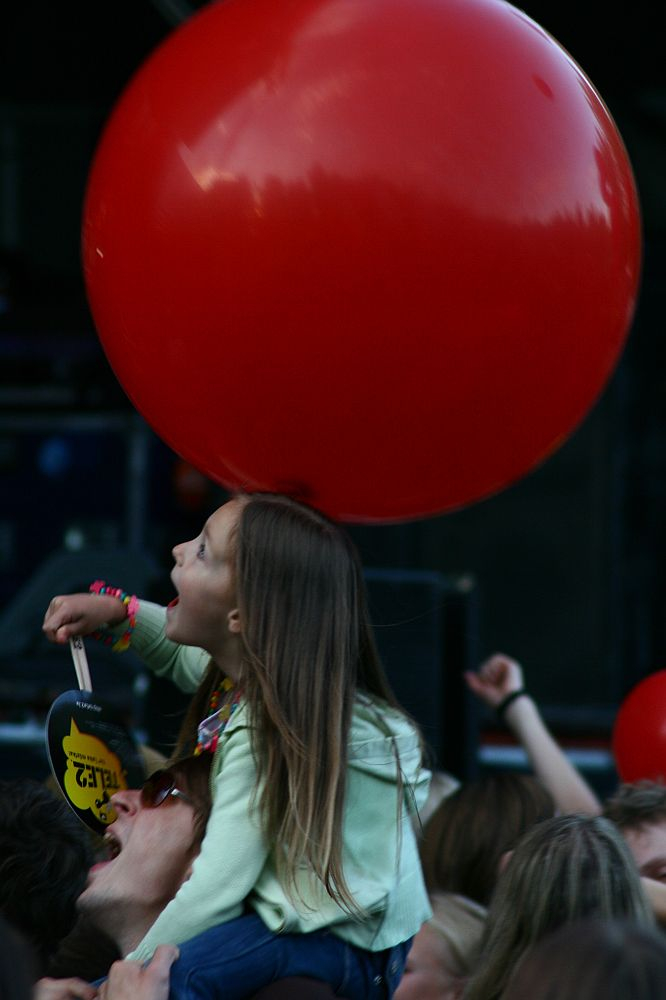 At Positivus, a young child with long hair, wearing a light green sweater, sits on an adult's shoulders in a crowd. The child looks up excitedly at a large red balloon above their head. They hold a small, round yellow sign. The background is dark, with blurred elements suggesting the lively atmosphere of AB 2008.