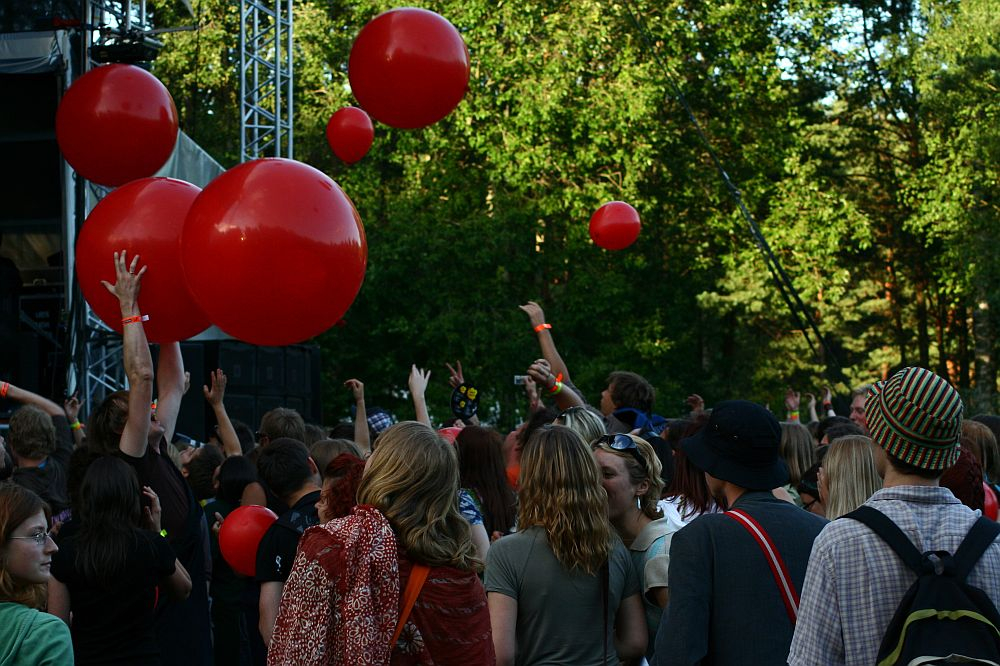 A crowd of people at the Positivus 2008 outdoor concert is enjoying the event. Large red balloons are being tossed among the attendees. The background is filled with lush green trees, and people are dressed in casual clothing. The atmosphere appears lively and festive.