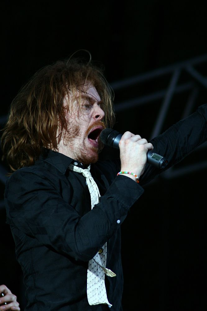 A singer with long, tousled hair passionately performs on stage at Positivus 2008, holding a microphone close to his mouth. He is wearing a black shirt and a light-colored tie. His right arm is raised, and his face is animated with emotion. The background is dark with part of a metal structure visible.