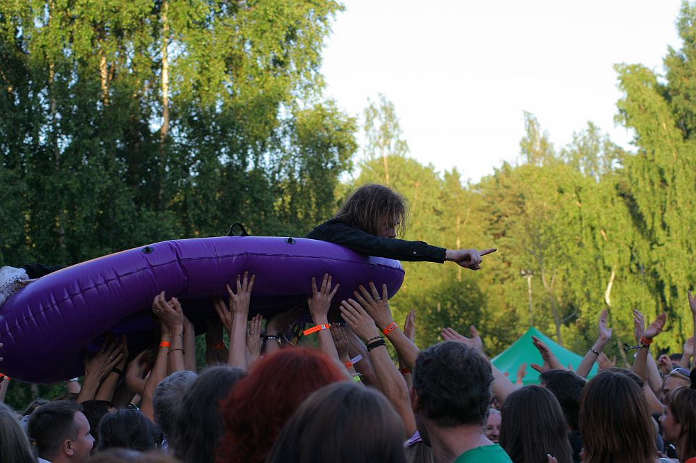 A crowd of people, many with raised hands, holds up a person riding a purple inflatable raft. The individual on the raft points outward with their right hand. The outdoor setting, surrounded by trees and vibrant foliage, hints at the lively atmosphere of an event like Positivus or AB 2008.