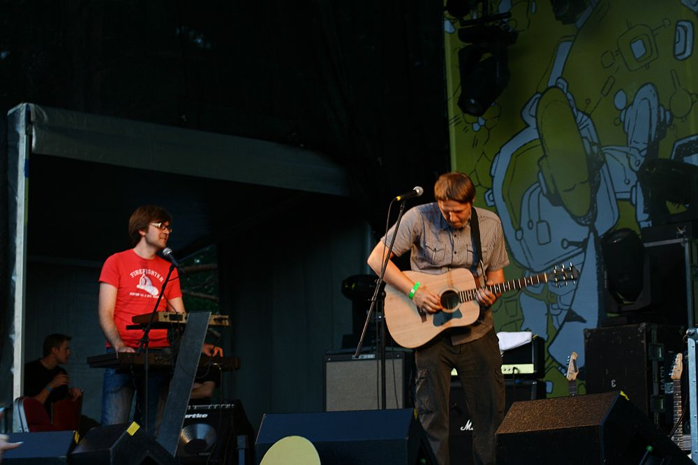 Two musicians perform on an outdoor stage at AB 2008 Positivus. The musician on the right plays an acoustic guitar and sings into a microphone, while the musician on the left, in a red shirt with glasses, plays a keyboard. Equipment and speakers surround them against a backdrop of bright, abstract design.