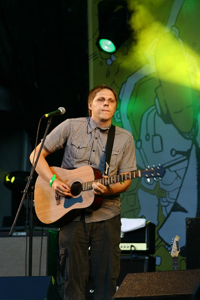 A person stands on an outdoor stage at AB 2008, playing an acoustic guitar. They are wearing a checked shirt and cargo pants. Behind them, a yellow and green abstract backdrop is illuminated by stage lights. A microphone stand is positioned in front of the person, and audio equipment is visible in the background.