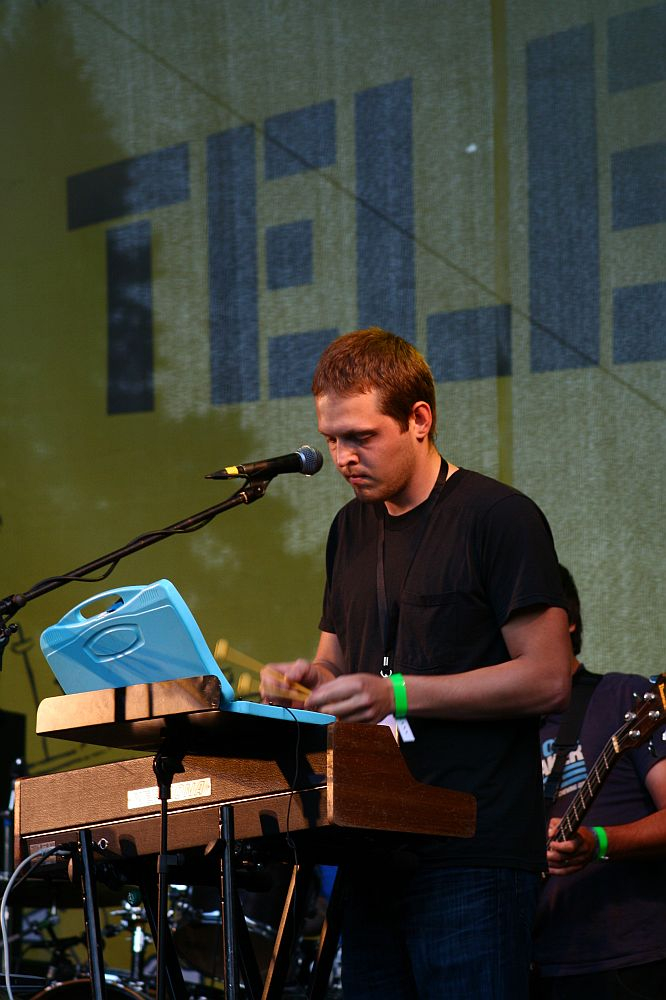 A man standing and playing a keyboard on a wooden stand under a microphone at Positivus. Wearing a black shirt and a wristband on his left hand, he captivates the crowd. In the background, an obscured band member plays guitar, with "TELE" visible on the 2008 backdrop behind them.
