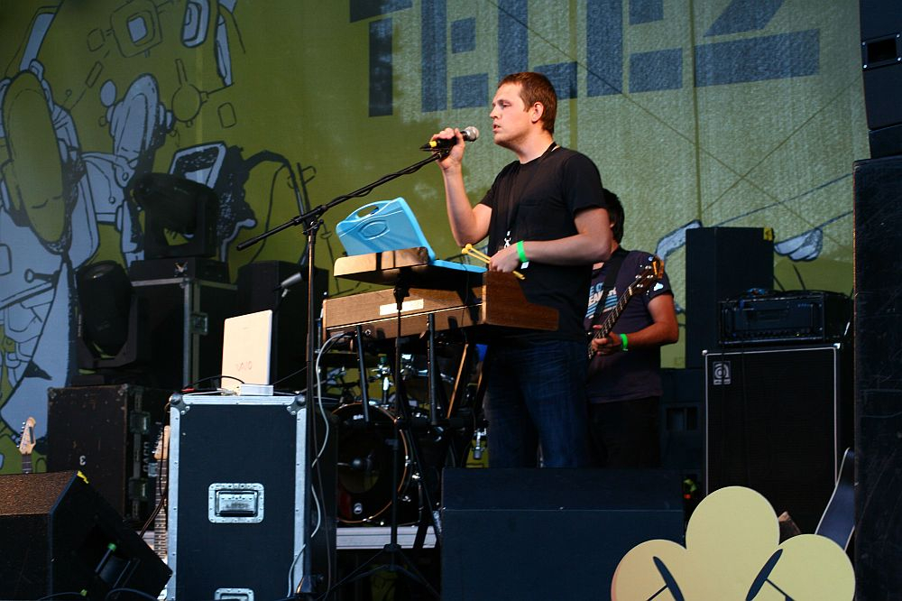 Man in black t-shirt sings into microphone while standing behind keyboard and computer on stage at Positivus. Another person in a patterned shirt plays guitar in background. Yellow and green backdrop with abstract designs and partial text "TELJ" visible. Various audio equipment surrounds the performers, reminiscent of AB 2008 vibes.