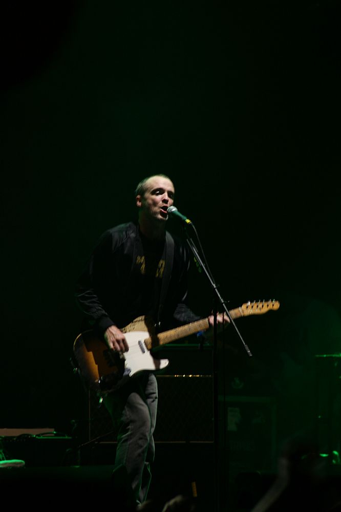 In a dimly lit environment, a person performs on stage at Positivus 2008, playing an electric guitar and singing into a microphone. The performer is dressed in casual clothing, including a dark jacket. The background is mostly dark, creating a moody atmosphere, with only a few details visible in the shadows.