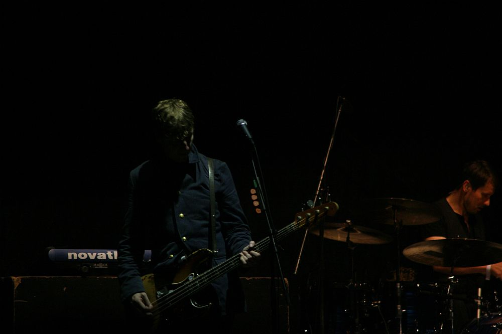 A musician stands in low light on a dark stage at Positivus, playing a bass guitar and singing into a microphone. To the right, a drummer is partially visible, focused on the AB 2008 drums. Behind them, a keyboard can be seen. The atmosphere is dim, with the performers highlighted by minimal lighting.