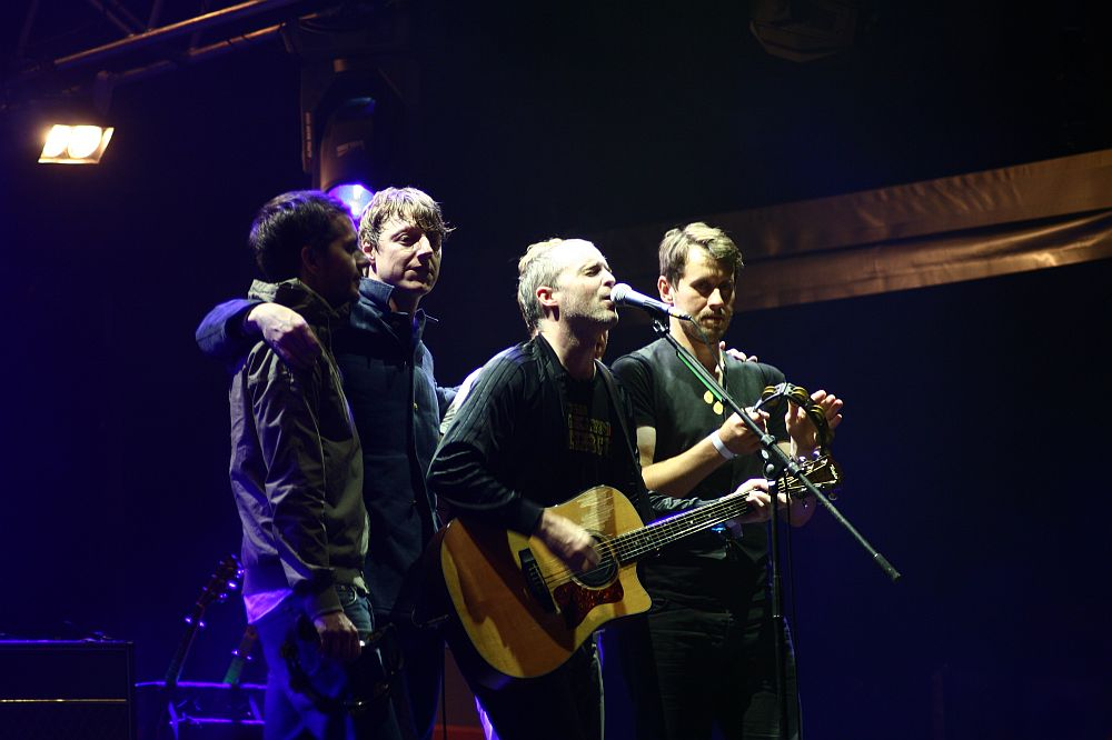 Four musicians perform on stage under dim, dramatic lighting at AB Positivus 2008. The man in the center sings into a microphone while playing an acoustic guitar. Another man plays a tambourine, and the two others stand close, engaging with the performance and the crowd. Stage equipment and lights are visible in the background.